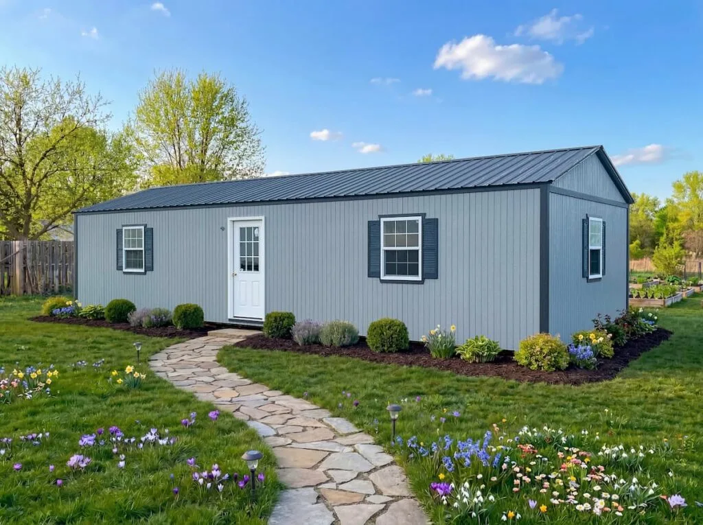 Elite Cabin with gray siding, black metal roof, white door, windows, shutters, and stone walkway in a landscaped yard.
