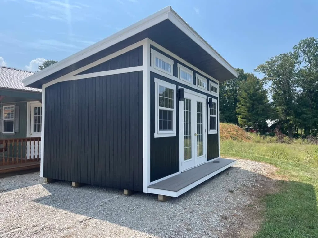 Black Elite She Shed Studio with white trim, gable roof accent, French doors, and a composite porch on a gravel pad in a rural setting.