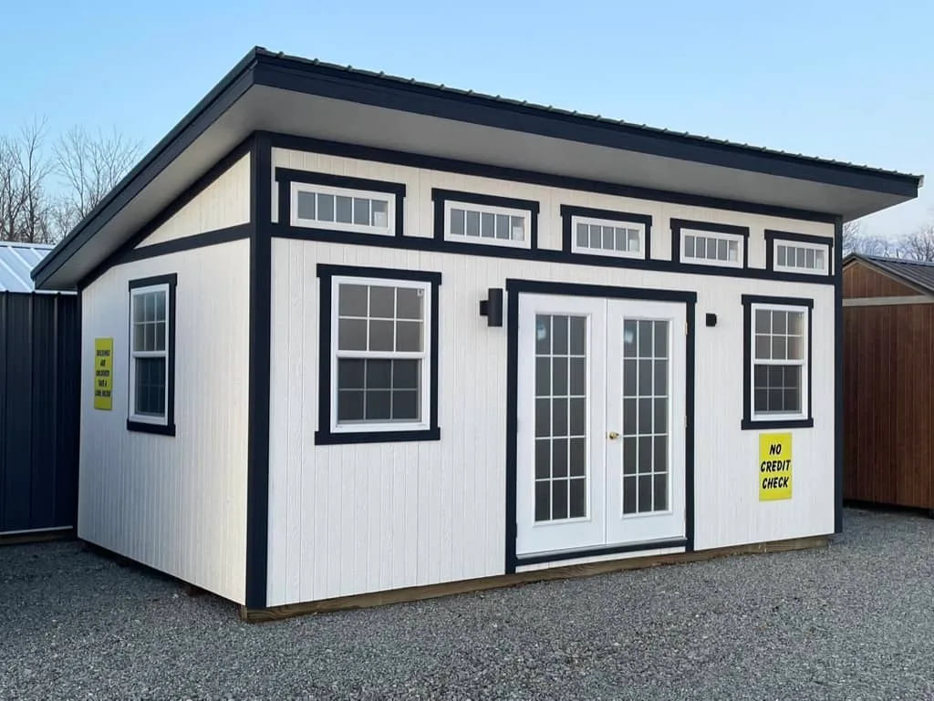 White Elite She Shed Studio with black trim, French doors, and a row of clerestory windows on a gravel sales lot.